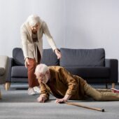 Senior Woman Helping To Get Up Fallen Husband Lying On Floor Near Walking Stick