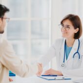 Friendly Female Doctor Greets Patient With Handshake, Pose In Private Clinic, Sit Opposite Each Other, Have Talk, Agree To Sign Insurance Contract, Meet In Hospital. Health Care And Medicine Concept
